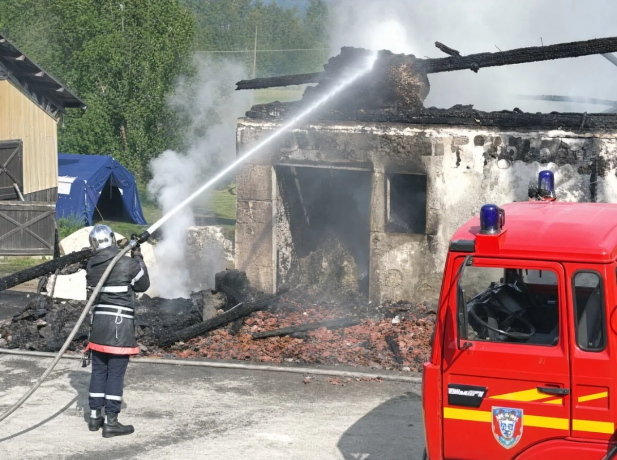 Sapeur-pompier projetant une lance à eau sur les décombres calcinés du centre équestre, fourgon d'incendie au premier plan